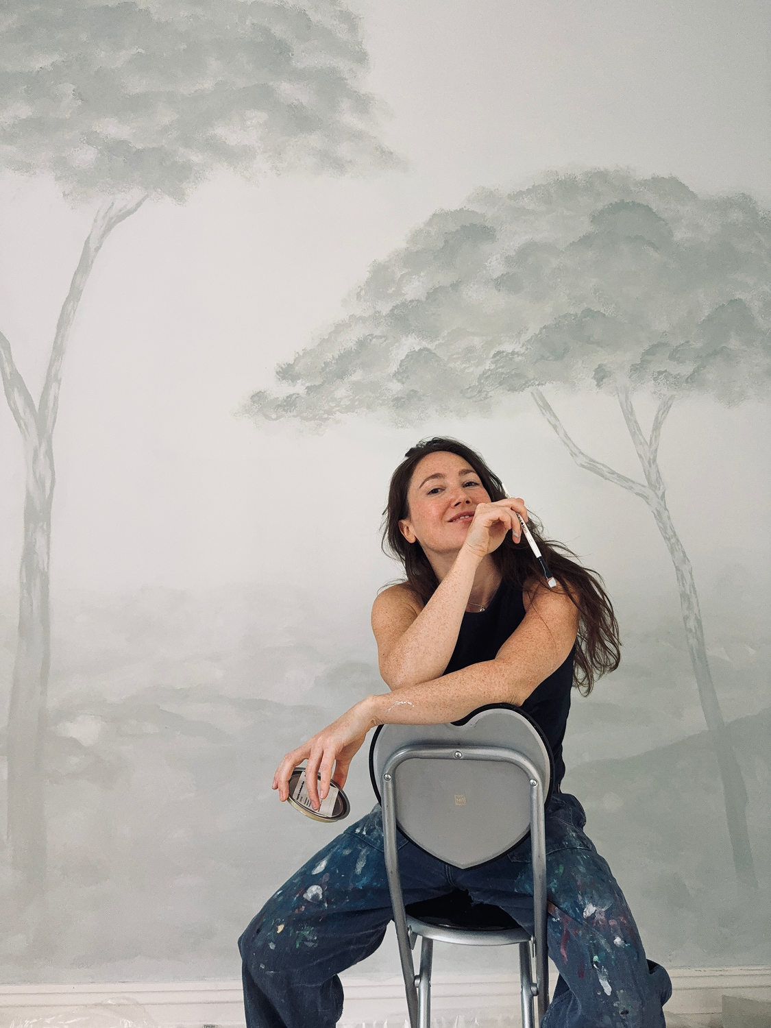 Artist sitting in front of ethereal tree mural in soft gray-green tones